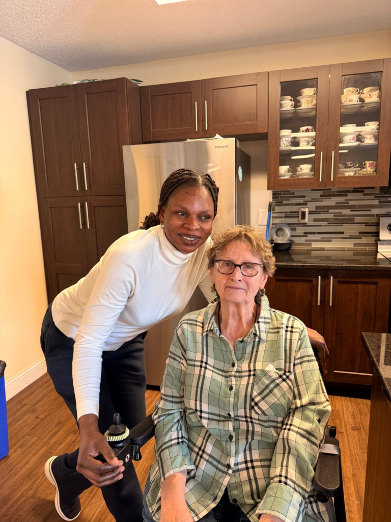 Tinalas caregiver smiling beside a client seated in a wheelchair in her kitchen